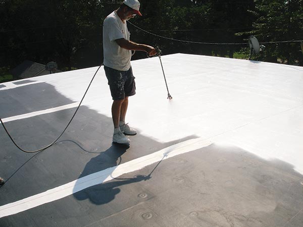 Flat-Roof-Coatings-Van-Nuys-CA-California-1 | Shark Roofing Systems Man wearing a hat and white shirt stands on a flat white roof, rolling roof coating with a long-handled roller.