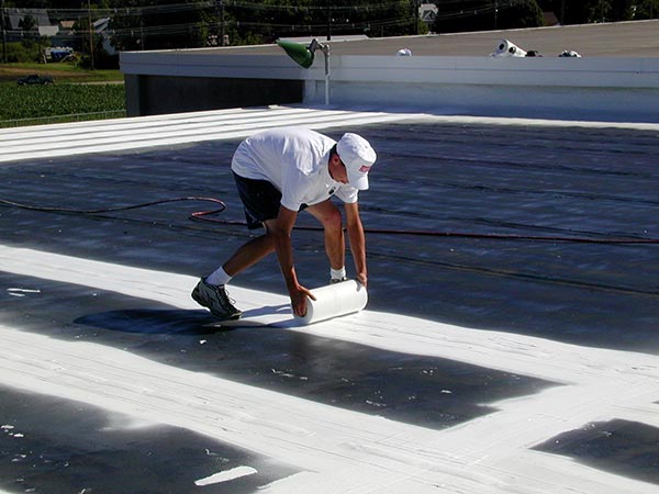 Flat-Roof-Coatings-Van-Nuys-CA-California-2 | Shark Roofing Systems A worker in a white shirt and cap unrolling white roofing membrane on a flat roof.