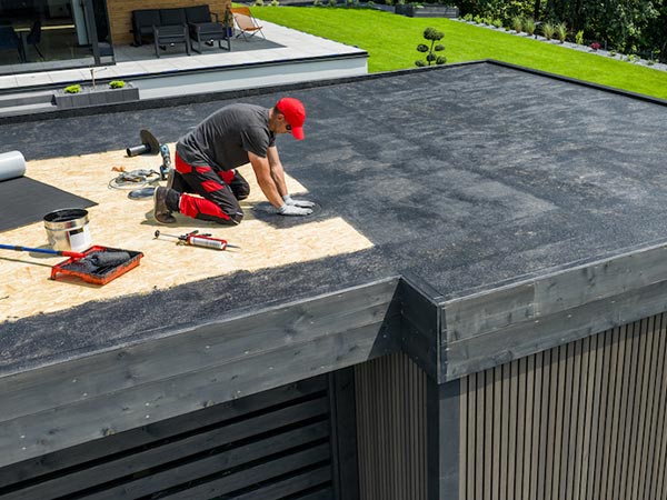 Roof-Maintenance-Glendale-CA-california-2 | Shark Roofing Systems Roofer in a red cap and gray shirt kneels on a flat roof, smoothing roofing material with a trowel while tools and a bucket sit nearby.