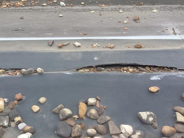 flat-roof-repair-Glendale-CA-California-1 | Shark Roofing Systems Close-up of cracked asphalt with a long gap and scattered rocks along a road edge/shoulder of a parking lot or roadway surface.