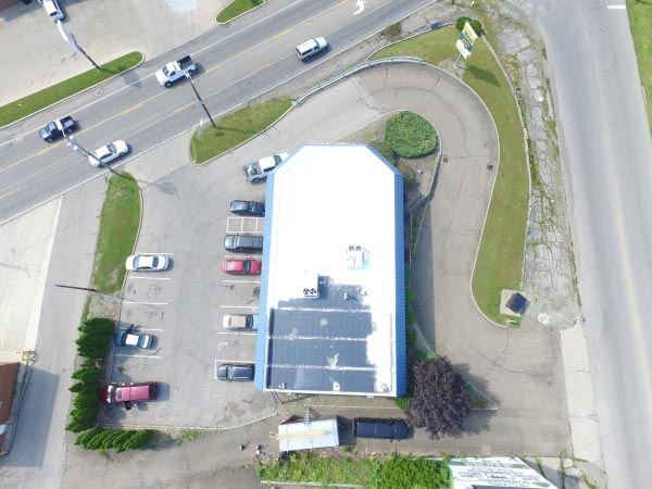 roof-restoration-Glendale-CA-California-1 | Shark Roofing Systems Aerial view of a blue-roofed rectangular building with a parking lot and curved driveway surrounded by green grass and shrubs.