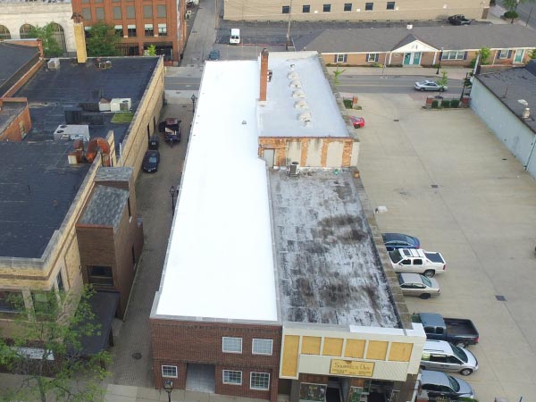 roof-restoration-Glendale-CA-California-2 | Shark Roofing Systems Aerial view of a row of brick commercial buildings with a bright white flat roof on the left and a darker, weathered roof on the right; a parking area with several cars is visible to the far right, and surrounding streets and smaller buildings frame the scene.
